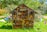 A wooden insect hotel stands in a lush green garden, filled with a variety of natural materials like logs, sticks, pine cones, and clay pots. It has a pitched roof and is surrounded by colorful triangular bunting, creating a festive atmosphere. The structure is set against a backdrop of dense greenery under a bright blue sky.