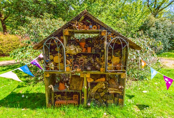 A wooden insect hotel stands in a lush green garden, filled with a variety of natural materials like logs, sticks, pine cones, and clay pots. It has a pitched roof and is surrounded by colorful triangular bunting, creating a festive atmosphere. The structure is set against a backdrop of dense greenery under a bright blue sky.