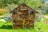 A wooden insect hotel stands in a lush green garden, filled with a variety of natural materials like logs, sticks, pine cones, and clay pots. It has a pitched roof and is surrounded by colorful triangular bunting, creating a festive atmosphere. The structure is set against a backdrop of dense greenery under a bright blue sky.