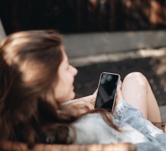 woman in white tank top lying on bed using black smartphone