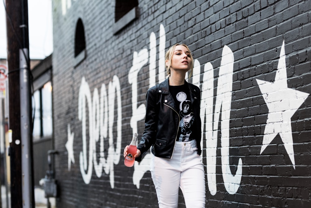 woman in black leather jacket and white pants standing beside brick wall during daytime