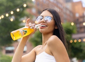 A woman with long brown hair and wearing sunglasses smiles while holding a bottle of Jarritos soda. She is outdoors, with blurred string lights and greenery in the background. Her nails are painted turquoise, and she is dressed in a white tank top.