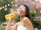 A woman with long brown hair and wearing sunglasses smiles while holding a bottle of Jarritos soda. She is outdoors, with blurred string lights and greenery in the background. Her nails are painted turquoise, and she is dressed in a white tank top.