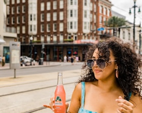 A woman with curly hair wearing sunglasses and a blue dress is holding a bottle of Jarritos guava-flavored soda. She is standing in an urban setting with a multistory brick building visible in the background along with a few people walking on the street.