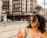 A woman with curly hair wearing sunglasses and a blue dress is holding a bottle of Jarritos guava-flavored soda. She is standing in an urban setting with a multistory brick building visible in the background along with a few people walking on the street.