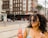 A woman with curly hair wearing sunglasses and a blue dress is holding a bottle of Jarritos guava-flavored soda. She is standing in an urban setting with a multistory brick building visible in the background along with a few people walking on the street.