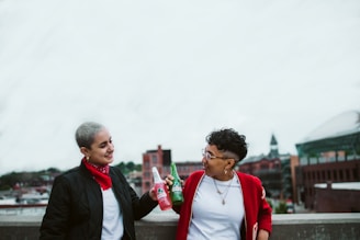 Two people stand on a rooftop with an urban background, each holding a bottle of drink and sharing a toast. One is wearing a black jacket over a white shirt and a red bandana around their neck, while the other is wearing a red jacket over a white shirt. Both are smiling and looking at each other.