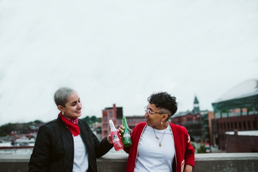 Two people stand on a rooftop with an urban background, each holding a bottle of drink and sharing a toast. One is wearing a black jacket over a white shirt and a red bandana around their neck, while the other is wearing a red jacket over a white shirt. Both are smiling and looking at each other.