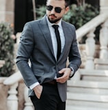 man in gray suit jacket and black pants wearing black sunglasses standing on gray concrete stairs