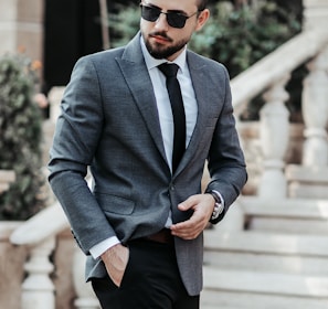 man in gray suit jacket and black pants wearing black sunglasses standing on gray concrete stairs