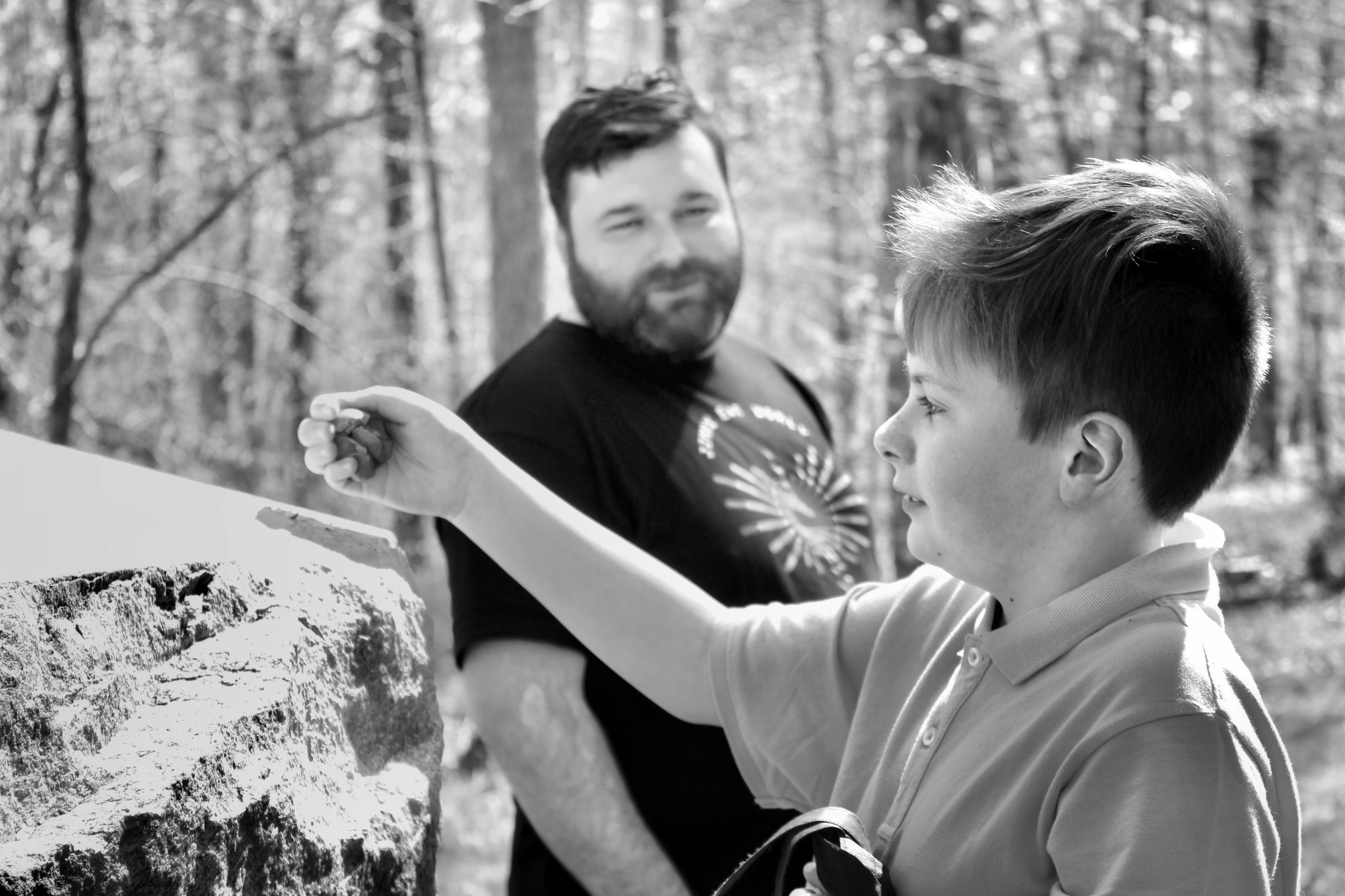 Photo: A boy extends his arm toward a rough stone wall while a bearded man watches from behind in a sunlit forest.