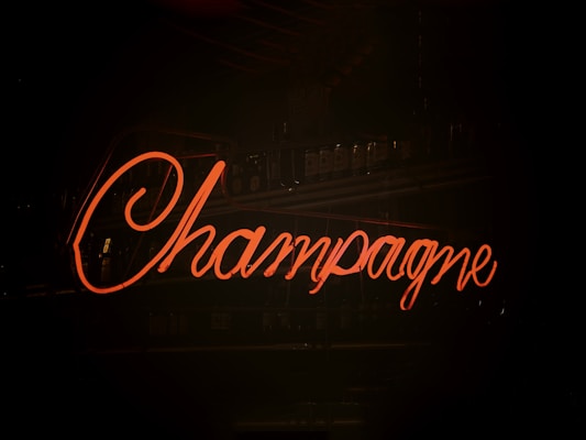 Neon sign with the word 'Champagne' glowing in a cursive font against a dark background, suggesting a bar or nightclub setting.