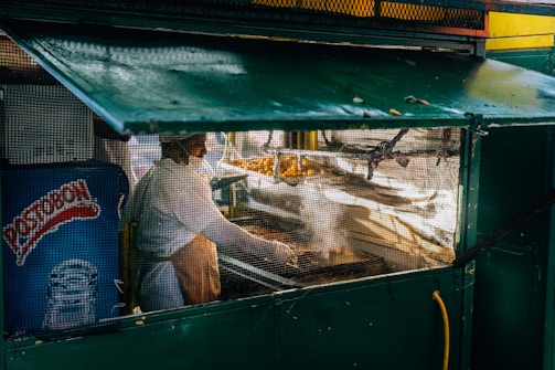 A street vendor is cooking under a green awning, behind a mesh screen. The vendor is wearing a white shirt and an apron. Next to them, there is a fridge with a 'Postobon' logo on it. The scene suggests an outdoor setting with cooking equipment and some food items visible.