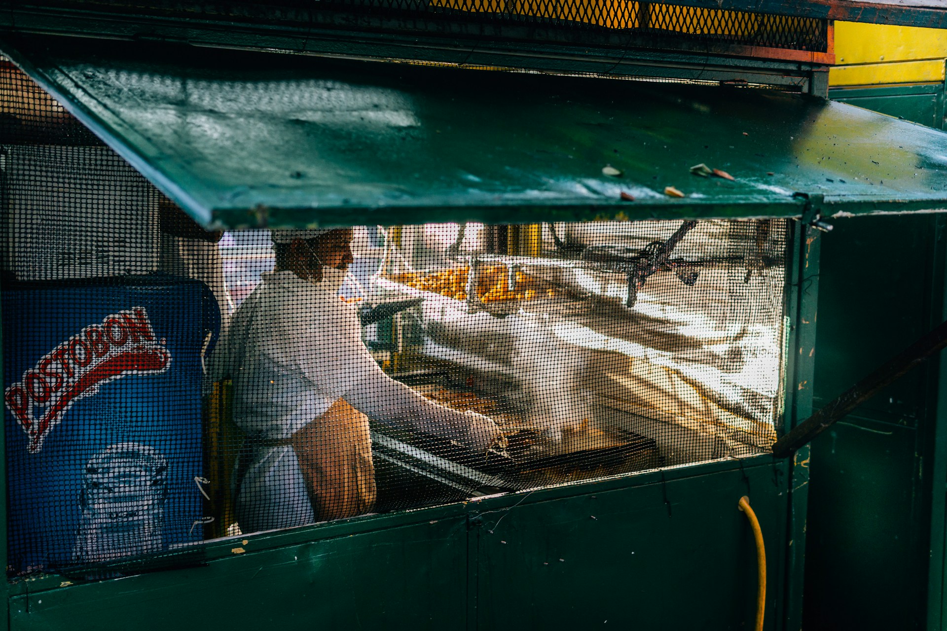 A street vendor is cooking under a green awning, behind a mesh screen. The vendor is wearing a white shirt and an apron. Next to them, there is a fridge with a 'Postobon' logo on it. The scene suggests an outdoor setting with cooking equipment and some food items visible.