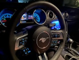 The interior of a car focusing on the steering wheel, which features a prominent horse emblem. The dashboard displays illuminated gauges and control buttons. Blue ambient lighting highlights various controls and dials.