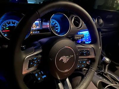 The interior of a car focusing on the steering wheel, which features a prominent horse emblem. The dashboard displays illuminated gauges and control buttons. Blue ambient lighting highlights various controls and dials.