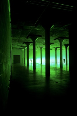 A large, dimly lit industrial space featuring tall columns casting shadows. Two people are silhouetted against a bright, green-lit wall at the far end, creating an eerie and mysterious atmosphere.