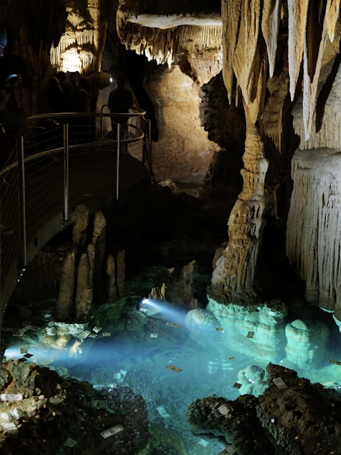 Intricate stalactites and stalagmites are illuminated in a dimly lit cave, creating a mystical atmosphere. A pool of clear water reflects the formations, while scattered currency notes are visible within the water. A railing allows visitors to safely view the natural formations.