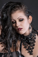 A person with Gothic style makeup, including dramatic eyeliner and red lipstick, is shown with closed eyes. Dark, wavy hair falls over the shoulder, and they wear elaborate black jewelry featuring beads and large stones, paired with matching earrings. A nose ring and lip piercing are also visible.