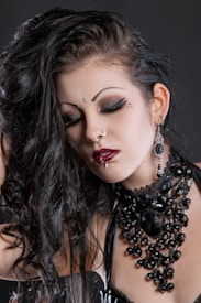 A person with Gothic style makeup, including dramatic eyeliner and red lipstick, is shown with closed eyes. Dark, wavy hair falls over the shoulder, and they wear elaborate black jewelry featuring beads and large stones, paired with matching earrings. A nose ring and lip piercing are also visible.