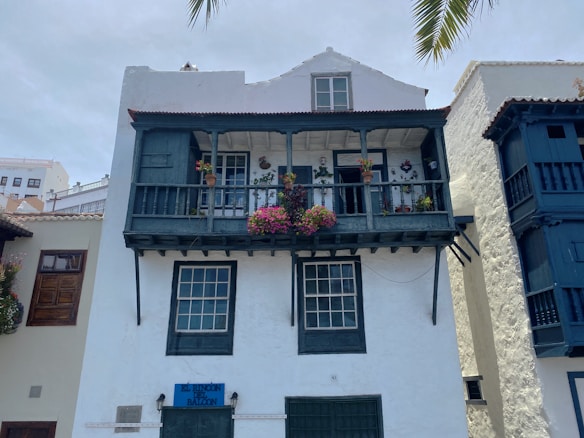 A traditional building with a wooden balcony featuring multiple potted plants, including vibrant flowers. The structure appears to have a rustic charm, with dark green shutters and windows. The blue sign reads 'El Rinc&oacute;n del Balc&oacute;n.' The surrounding architecture is in a similar style, with white walls and dark wooden elements.