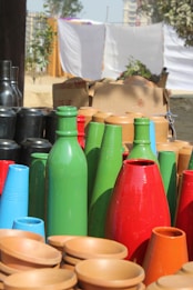 A collection of colorful pots and vases made of clay and painted in bright colors like green, red, blue, and black is displayed outdoors. The arrangement also includes unpainted terracotta bowls. In the background, a cardboard box and sheets hanging on a line are visible, with greenery and a building in the distance.