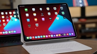 A tablet with a keyboard attachment is displayed on a wooden surface. The screen shows a home screen with multiple app icons arranged in rows against a red and blue background. In the background, part of another similar device is visible, reflecting the main screen.