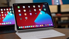 A tablet with a keyboard attachment is displayed on a wooden surface. The screen shows a home screen with multiple app icons arranged in rows against a red and blue background. In the background, part of another similar device is visible, reflecting the main screen.