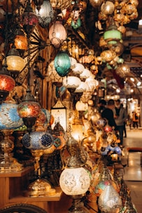 A collection of intricately designed mosaic lamps in various shapes and colors displayed on shelves. The lamps are adorned with vibrant patterns and are lit, casting a warm glow. The setting appears to be a bustling marketplace or bazaar with other decor items visible in the background.