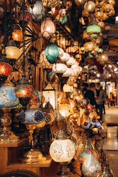 A collection of intricately designed mosaic lamps in various shapes and colors displayed on shelves. The lamps are adorned with vibrant patterns and are lit, casting a warm glow. The setting appears to be a bustling marketplace or bazaar with other decor items visible in the background.