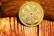 A gold-colored coin is placed against a background that appears to be wooden. The coin has intricate designs and the inscriptions 'CARDANO', 'SECURE LAYERED SMART CONTRACTS', and 'ADA' can be seen.
