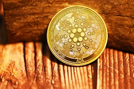 A gold-colored coin is placed against a background that appears to be wooden. The coin has intricate designs and the inscriptions 'CARDANO', 'SECURE LAYERED SMART CONTRACTS', and 'ADA' can be seen.
