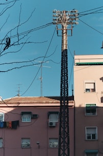 A tall utility pole with multiple power lines and cables extends above residential buildings. The buildings have a rustic, weathered appearance, with faded pink walls, satellite dishes, air conditioners, and laundry hanging out to dry. Leafless tree branches are in the foreground, contrasting against a bright, clear blue sky.