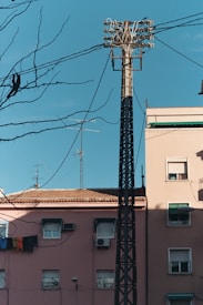 A tall utility pole with multiple power lines and cables extends above residential buildings. The buildings have a rustic, weathered appearance, with faded pink walls, satellite dishes, air conditioners, and laundry hanging out to dry. Leafless tree branches are in the foreground, contrasting against a bright, clear blue sky.