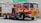 A red fire truck is parked outside a Safeway store. The truck is a Scania model, marked with 'Metropolitan Fire Brigade' and equipped with water hoses and rescue gear. Bright safety stripes and emergency lights are visible, emphasizing its readiness for action.