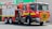 A red fire truck is parked outside a Safeway store. The truck is a Scania model, marked with 'Metropolitan Fire Brigade' and equipped with water hoses and rescue gear. Bright safety stripes and emergency lights are visible, emphasizing its readiness for action.