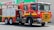 A red fire truck is parked outside a Safeway store. The truck is a Scania model, marked with 'Metropolitan Fire Brigade' and equipped with water hoses and rescue gear. Bright safety stripes and emergency lights are visible, emphasizing its readiness for action.