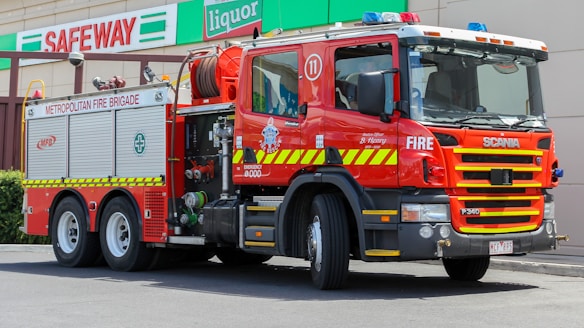 A red fire truck is parked outside a Safeway store. The truck is a Scania model, marked with 'Metropolitan Fire Brigade' and equipped with water hoses and rescue gear. Bright safety stripes and emergency lights are visible, emphasizing its readiness for action.