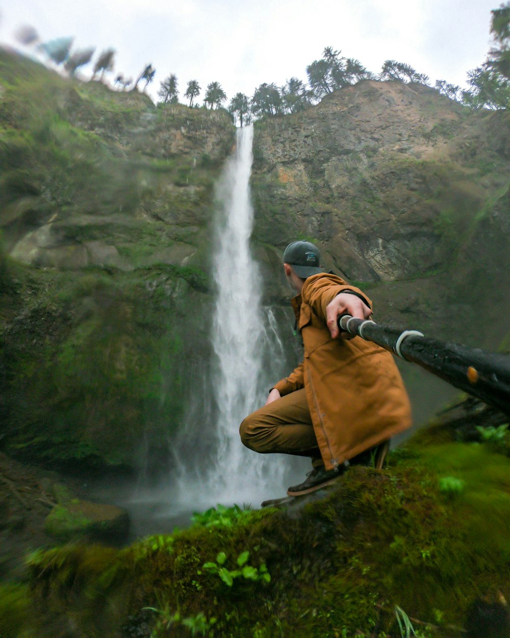 man in brown jacket and black pants standing on green grass near waterfalls during daytime