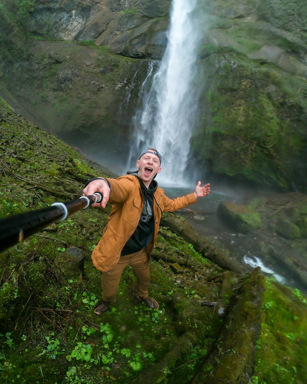 man in orange jacket and brown pants standing on green grass field near waterfalls during daytime