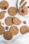 Several round, brown cookies with visible oatmeal flakes are arranged on a white surface. Some of the cookies are whole, while one is partially broken. Star anise and cinnamon sticks are placed around the cookies for garnish, adding a rustic decorative touch.