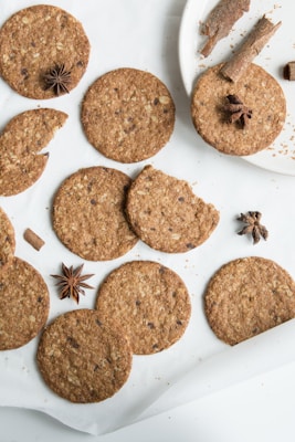 Several round, brown cookies with visible oatmeal flakes are arranged on a white surface. Some of the cookies are whole, while one is partially broken. Star anise and cinnamon sticks are placed around the cookies for garnish, adding a rustic decorative touch.