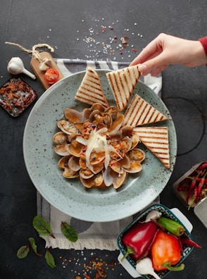 A gray speckled plate is filled with a dish of clams cooked in sauce, topped with shreds of cheese and garnished with red pepper flakes. Triangular pieces of grilled bread are arranged around the clams. A hand is seen reaching to take a piece of bread. The setting includes ingredients such as whole garlic, cherry tomatoes, and various peppers placed nearby on a dark countertop. A small basket containing red, orange, and green peppers is also visible.