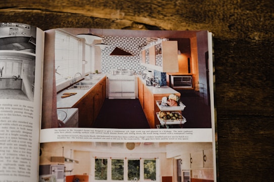 A vintage kitchen design is featured in a magazine spread. The kitchen includes wooden cabinetry, white countertops, and a tiled backsplash with a geometric pattern. Various kitchen appliances such as a toaster oven and a sink with a faucet are visible. Fresh produce is placed in baskets on a lower shelf, and natural light enters through a large window.