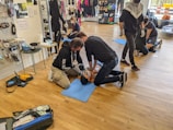 People are practicing CPR techniques on medical mannequins in a room filled with various equipment and supplies. Several individuals are kneeling on mats placed on a wooden floor, performing chest compressions and rescue breaths. There are medical kits and defibrillator equipment visible, and some standing individuals observing or preparing to assist.