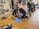 People are practicing CPR techniques on medical mannequins in a room filled with various equipment and supplies. Several individuals are kneeling on mats placed on a wooden floor, performing chest compressions and rescue breaths. There are medical kits and defibrillator equipment visible, and some standing individuals observing or preparing to assist.