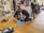 People are practicing CPR techniques on medical mannequins in a room filled with various equipment and supplies. Several individuals are kneeling on mats placed on a wooden floor, performing chest compressions and rescue breaths. There are medical kits and defibrillator equipment visible, and some standing individuals observing or preparing to assist.