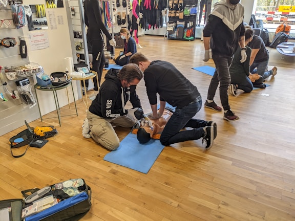 People are practicing CPR techniques on medical mannequins in a room filled with various equipment and supplies. Several individuals are kneeling on mats placed on a wooden floor, performing chest compressions and rescue breaths. There are medical kits and defibrillator equipment visible, and some standing individuals observing or preparing to assist.