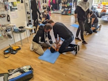 People are practicing CPR techniques on medical mannequins in a room filled with various equipment and supplies. Several individuals are kneeling on mats placed on a wooden floor, performing chest compressions and rescue breaths. There are medical kits and defibrillator equipment visible, and some standing individuals observing or preparing to assist.