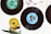 Vinyl records are splayed out on a white surface, with labels in different colors, such as green and blue. A skateboard wheel and truck, positioned in the lower left corner, add an eclectic touch to the arrangement. The top portion of a record sleeve is visible in the corner, featuring printed text and abstract artwork.